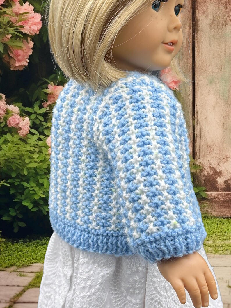 May include: A blue and white crochet sweater on a doll. The sweater has a textured pattern and a ribbed hem and cuffs. The doll is wearing a white dress. The background includes pink flowers and a wooden fence.
