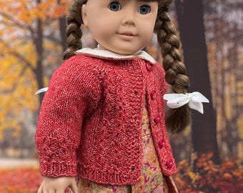 May include: A doll with brown hair in two braids, wearing a red knit cardigan with a white collar over a floral print dress. The cardigan features a textured cable knit pattern and red buttons. The doll has blue eyes and white hair ribbons.