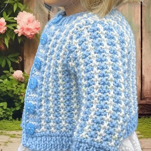 May include: A blue and white knitted cardigan with flower-shaped buttons. The cardigan has a textured pattern and a ribbed hem and cuffs. The garment is designed for a child and is worn over a white dress. The background includes pink flowers and a wooden fence.