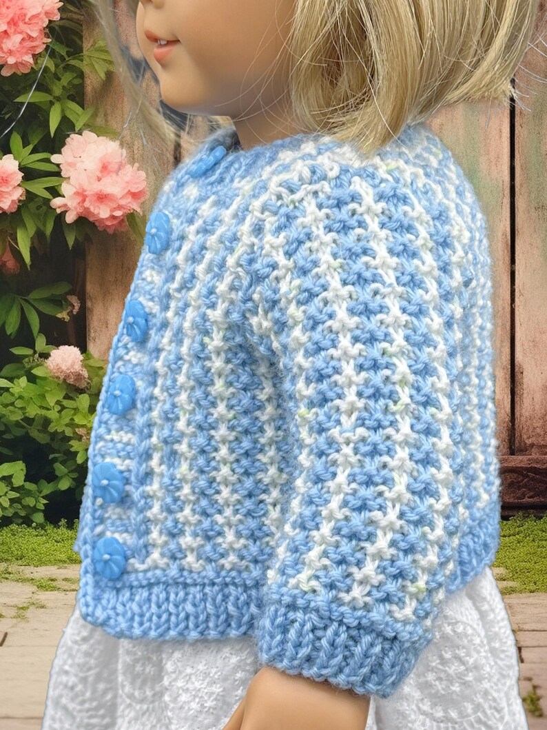 May include: A blue and white knitted cardigan with flower-shaped buttons. The cardigan has a textured pattern and a ribbed hem and cuffs. The garment is designed for a child and is worn over a white dress. The background includes pink flowers and a wooden fence.