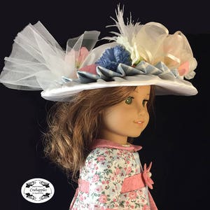 May include: A white satin doll hat with blue and white gingham ribbon, white tulle, and pink and blue flowers. The hat is worn by a doll with brown hair and green eyes.