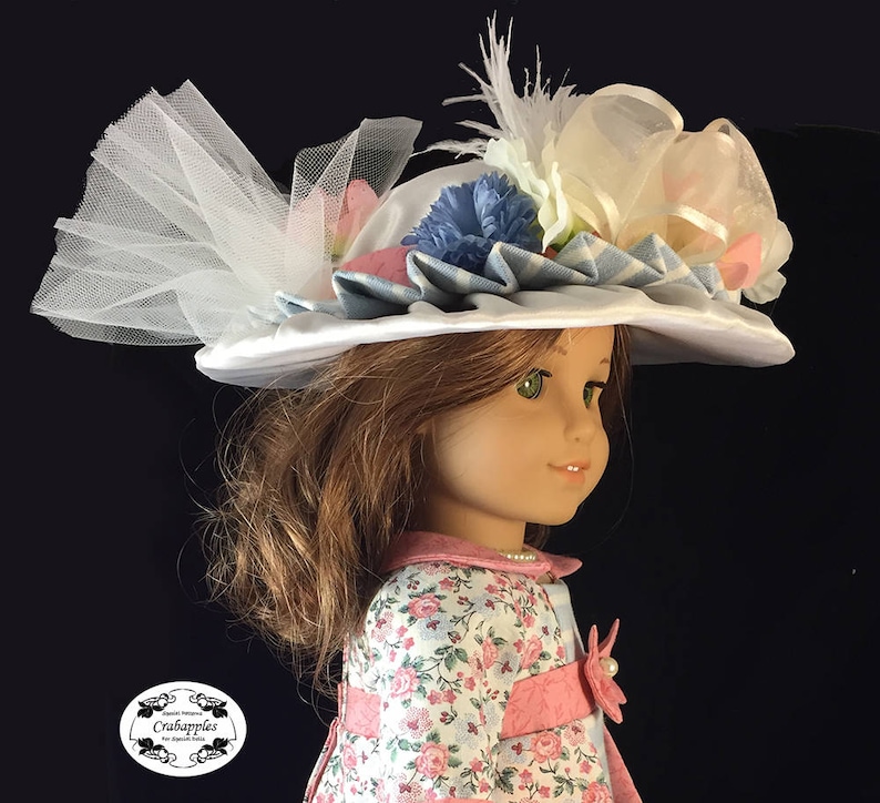 May include: A white satin doll hat with blue and white gingham ribbon, white tulle, and pink and blue flowers. The hat is worn by a doll with brown hair and green eyes.