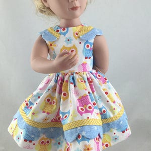 May include: A doll wearing a blue and yellow dress with a pattern of owls and flowers. The dress has a scalloped hem and a Peter Pan collar. The doll is wearing blue shoes.