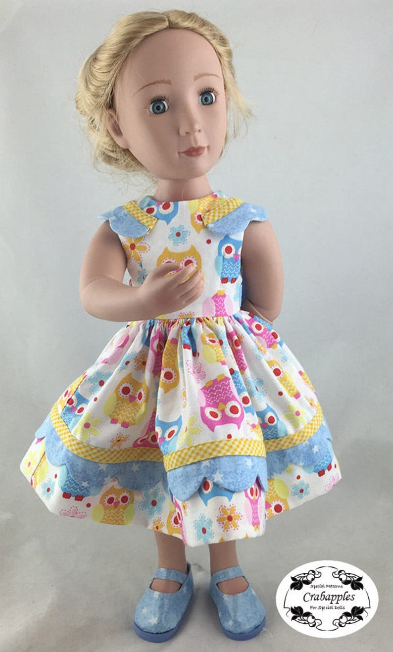 May include: A doll wearing a blue and yellow dress with a pattern of owls and flowers. The dress has a scalloped hem and a Peter Pan collar. The doll is wearing blue shoes.