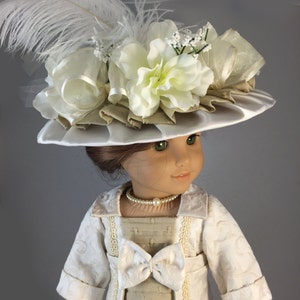 May include: A doll wearing a white dress with a beige jacket and a white hat with a large white flower and a white feather.