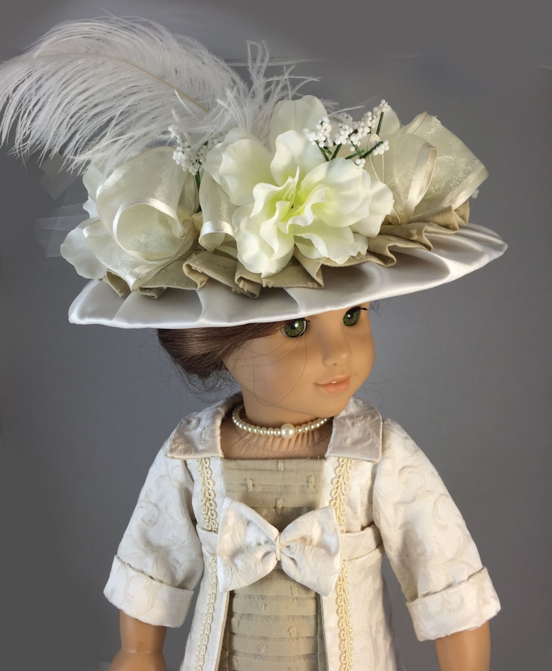 May include: A doll wearing a white dress with a beige jacket and a white hat with a large white flower and a white feather.