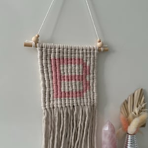 Könnte beinhalten: Beige Makramee-Wandbehang mit dem Wort "LOVE" in Rosa. Das Makramee ist an einem Holzdübel mit Holzperlen aufgehängt. Der Wandbehang hat unten eine Fransen. Ein Rosenquarz-Kristall und eine dekorative Vase sind ebenfalls im Bild.