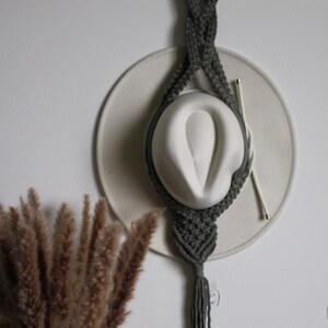 Op de afbeelding: Een grijze macramé hoedenhouder met een houten ring, met een witte hoed. De houder hangt aan een witte muur. Gedroogde bruine decoratieve planten staan in de linker benedenhoek.