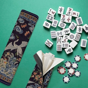 Könnte beinhalten: Ein komplettes Mahjong-Set mit weißen Spielsteinen mit chinesischen Schriftzeichen und Symbolen sowie bunten Chips. Das Set enthält eine marineblaue Hülle mit Kranich- und Blumendesign, vor grünem Hintergrund.