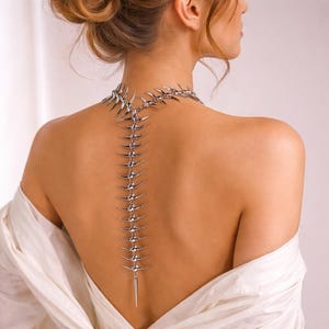 Puede incluir: Un collar plateado con un diseño en forma de espina dorsal, con una serie de elementos puntiagudos en la espalda. El collar se lleva en la espalda de una persona, con tela blanca sobre los hombros.