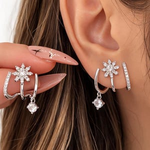 Könnte beinhalten: Silberne Ohrringe mit einem Schneeflocken-Design und funkelnden Kristallen. Die Ohrringe haben einen hängenden runden Kristall und einen gebogenen Stab mit eingebetteten Kristallen. Ein zweites Ohrring-Set enthält einen kristallbesetzten Stab.