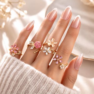 Könnte beinhalten: Nahaufnahme einer Hand mit mehreren goldfarbenen Ringen, jeweils mit floralem Design. Die Ringe haben rosa, weiße und klare Edelsteine. Die Hand trägt einen cremefarbenen Pullover, im Hintergrund sind Blumen zu sehen.
