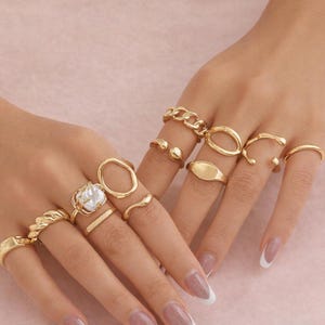 Könnte beinhalten: Auf zwei Händen sind goldfarbene Ringe in verschiedenen Designs zu sehen. Dazu gehören ein Kettenglied, ein Siegelring und ein Ring mit einem perlenartigen Stein. Der Hintergrund ist ein zartes Rosa.