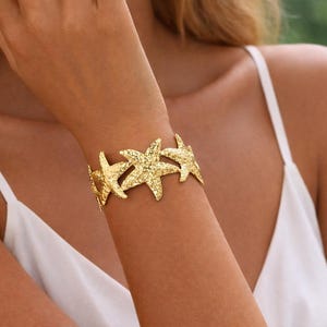 Puede incluir: Un brazalete dorado con forma de estrella de mar. El brazalete presenta múltiples diseños de estrellas de mar texturizadas unidas entre sí. La joya se lleva en la muñeca de una persona que lleva una camiseta blanca. El brazalete es una pieza llamativa.