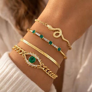 Puede incluir: Una colección de pulseras doradas en una muñeca. Las pulseras incluyen un diseño de serpiente, un estilo de cadena con una piedra de color esmeralda, una banda delgada con piedras de color esmeralda y un patrón de espiga.