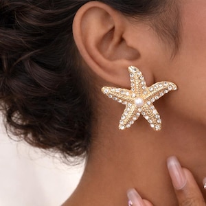 Può includere: Orecchino a forma di stella marina dorata con perla centrale e cristalli trasparenti. Il design a stella a cinque punte è un gioiello accattivante. L'orecchino singolo aggiunge un tocco di eleganza costiera a qualsiasi outfit.