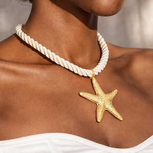 Può includere: Collana con pendente a forma di stella marina dorata e spessa catena a corda color crema. La stella marina è strutturata e ha una finitura metallica. La collana è indossata intorno al collo.