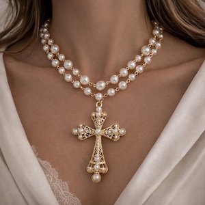 Puede incluir: Un collar con colgante de cruz dorada con una doble hilera de perlas de imitación. La cruz está adornada con perlas más pequeñas y detalles intrincados. El collar se lleva sobre una blusa de satén blanco.