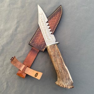 May include: A fixed-blade knife with a textured antler handle and a silver-colored metal blade featuring a serrated spine and etched script. The knife is paired with a brown leather sheath that has a stamped geometric pattern and a strap.