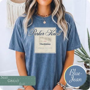 Può includere: Una t-shirt blu con lavaggio minerale caratterizzata da una grafica rettangolare sul petto. La grafica riporta il testo Parker Knoll, 1993, Chardonnay e Napa Valley, insieme a un disegno al tratto di un vigneto su sfondo color crema.