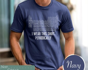 May include: A navy blue short-sleeved t-shirt featuring a white graphic of the periodic table of elements printed on the chest. Below the table, the text reads I WEAR THIS SHIRT PERIODICALLY in white capital letters. The shirt has a crew neckline.