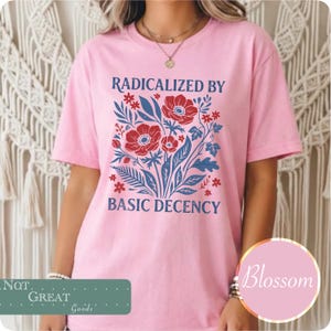 以下が含まれることがあります： 赤色の花と青色の葉のグラフィックが描かれたライトピンクのTシャツで、青色の文字で「RADICALIZED BY BASIC DECENCY」とプリントされています。デザインは胸の中央に配置されており、クルーネックと半袖が特徴です。
