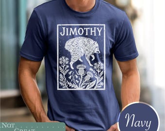 May include: A navy blue short-sleeve t-shirt featuring a white graphic print of a furry creature above a cluster of thistles and wildflowers. The text JIMOTHY is printed in a distressed white font at the top of the rectangular graphic design.