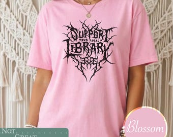 May include: A light pink short-sleeved t-shirt featuring a black graphic print on the chest. The design consists of intricate, spiky, metal-style lettering that reads "SUPPORT YOUR LOCAL LIBRARY" arranged in a symmetrical, thorn-like pattern.