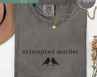 May include: A charcoal gray crew neck t-shirt featuring the text "attempted murder" in black lowercase letters above a graphic of two black crows facing each other. The shirt has a Comfort Colors label at the neck and a gold star pendant necklace.