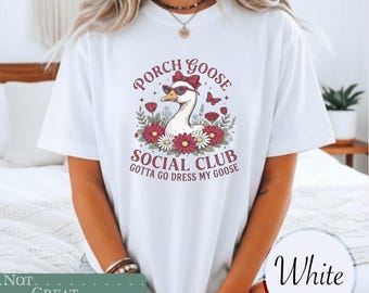 May include: A white short-sleeve t-shirt featuring a graphic of a goose wearing sunglasses and a red bow. The text reads PORCH GOOSE SOCIAL CLUB and GOTTA GO DRESS MY GOOSE, surrounded by red and white flowers and a small butterfly illustration.