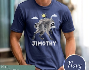 May include: A navy blue short-sleeve t-shirt featuring a graphic illustration of a furry raccoon walking under a yellow sun and white clouds. The text JIMOTHY is printed in white capital letters below the raccoon illustration on the chest.