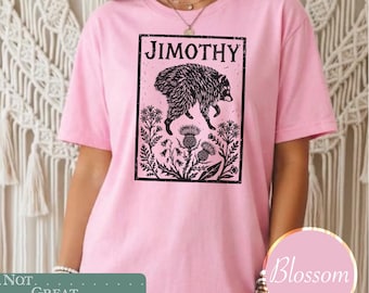 May include: A light pink short-sleeve t-shirt featuring a black rectangular graphic print. The design includes the text JIMOTHY above a detailed illustration of a raccoon surrounded by various botanical elements like thistles and wildflowers.