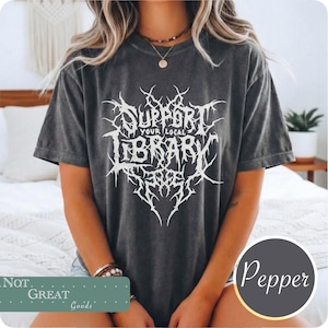 Puede incluir: Una camiseta de manga corta gris carbón con un estampado gráfico blanco en el pecho con el texto "SUPPORT YOUR LOCAL LIBRARY" en una fuente estilizada, dentada e inspirada en el metal. La prenda tiene cuello redondo y acabado desgastado.