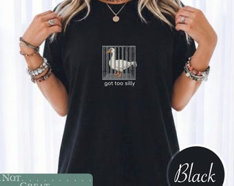 May include: A black short-sleeved t-shirt featuring a graphic of a white goose standing inside a gray metal cage. Below the cage, the text reads "got too silly" in a simple white font. The shirt has a crew neckline and a standard fit.