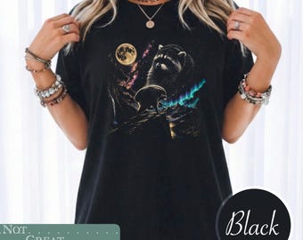 May include: A black t-shirt featuring a graphic print of a raccoon standing on a rocky ledge under a full moon and a vibrant aurora borealis. The design includes purple and blue nebula clouds in the night sky, creating a detailed celestial scene.