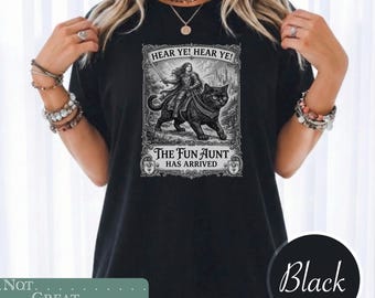 May include: A black t-shirt featuring a graphic print of a person in medieval armor riding a large black cat. The text reads HEAR YE! HEAR YE! THE FUN AUNT HAS ARRIVED. The design is enclosed in an ornate rectangular frame with a castle background.