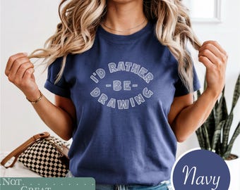 May include: A navy blue short-sleeve t-shirt featuring white text in a circular arrangement that reads I'D RATHER BE DRAWING. The lettering has a hatched, sketch-like texture. The shirt has a crew neckline and a standard fit for casual wear.