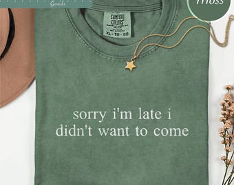 May include: A moss green short-sleeve t-shirt with a crew neckline and the text "sorry i'm late i didn't want to come" printed in white. The shirt features a Comfort Colors label at the inner neck and is paired with a gold star pendant necklace.