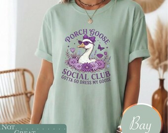 May include: A light green short-sleeve t-shirt featuring a graphic of a goose wearing purple sunglasses and a bow. The text reads Porch Goose Social Club and Gotta Go Dress My Goose, surrounded by purple flowers and a small butterfly illustration.