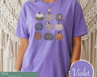 May include: A violet short-sleeved t-shirt featuring a graphic print of nine pumpkins arranged in a three-by-three grid. Each pumpkin displays a unique pattern, including floral, leopard print, star, and geometric designs in various muted colors.
