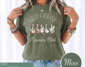 May include: A moss green short-sleeve t-shirt featuring a graphic of five white geese in various athletic poses. The text "SILLY GOOSE" appears above the geese, and "Running Club" is printed below them in a decorative, light-colored script font.