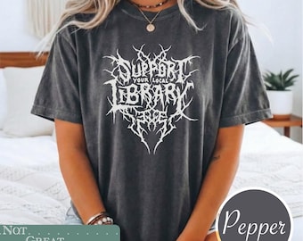 May include: A charcoal gray short-sleeve t-shirt featuring white graphic text that reads SUPPORT YOUR LOCAL LIBRARY in a stylized, jagged, metal-inspired font. The shirt has a washed, vintage-style finish and a classic crew neckline design.