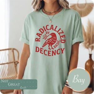 Op de afbeelding: Een lichtgroen t-shirt met korte mouwen en een rode afbeelding van een vogel die een takje vasthoudt. De tekst RADICALIZED BY BASIC DECENCY is in een cirkelvormig rood lettertype rond de vogel gedrukt. Het t-shirt heeft een ronde hals en een losse pasvorm.
