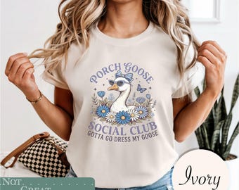 May include: An ivory t-shirt featuring a graphic of a goose wearing sunglasses and a blue bow. The text reads Porch Goose Social Club and Gotta Go Dress My Goose, surrounded by blue and white flowers and a small butterfly illustration.