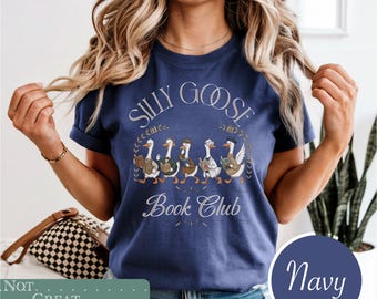 May include: A navy blue short-sleeve t-shirt featuring a graphic of five geese reading books. The text "Silly Goose Book Club" is printed in a light-colored font above and below the illustration of the geese wearing various hats and outfits.