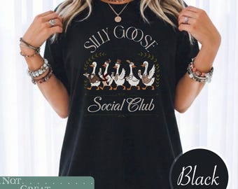 May include: A black t-shirt featuring a graphic of five geese dressed in formal attire, including hats and scarves. The text "SILLY GOOSE" appears above the geese, and "Social Club" is printed below them in a decorative, elegant script font.