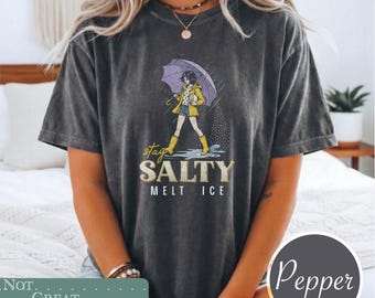 May include: A charcoal gray short-sleeve t-shirt featuring a graphic of a person in a yellow raincoat and boots holding a purple umbrella under rain. The text reads "stay SALTY MELT ICE" in a mix of script and block lettering centered on the chest.