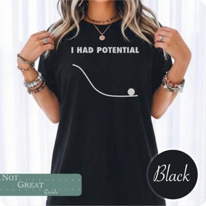 Peut inclure: Un t-shirt noir à manches courtes avec le texte blanc I HAD POTENTIAL au-dessus d'un graphique blanc représentant une ligne courbe en pente avec un petit cercle au bas, illustrant un concept physique d'énergie potentielle en design simple.