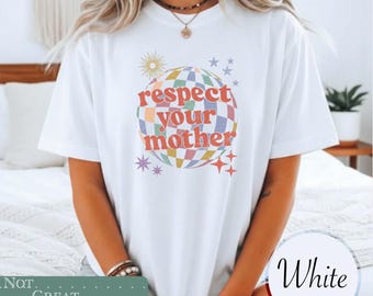 May include: A white short-sleeve t-shirt featuring a graphic of a multicolored checkered disco ball. The text "respect your mother" is printed in orange across the center of the disco ball, surrounded by small star and sun icons in various colors.