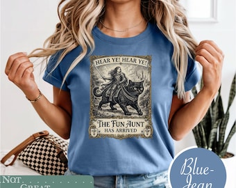 May include: A blue t-shirt featuring a graphic print of a person in armor riding a large black cat. The text reads HEAR YE! HEAR YE! THE FUN AUNT HAS ARRIVED. The design is framed in an ornate border with a castle in the background.